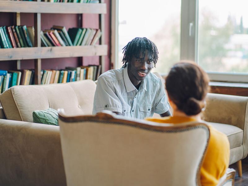 A young man sit on a sofa in an office speaking to another individual